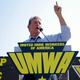 Sen. Joe Manchin standing in front of a United Mine Workers of America podium raising his left hand in the air.